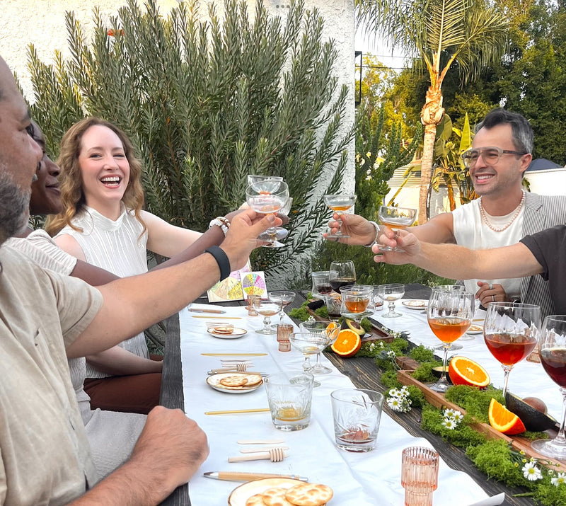 A group of friends and family enjoying a honey tasting experience together at an outdoor dinner table, raising honey-glasses in a toast. 