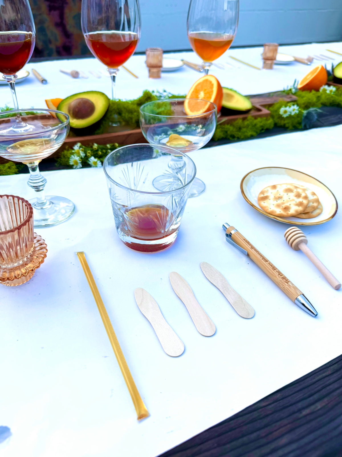 Formal  honey tasting table setting with clear glassware, garnished with citrus and avocado, alongside wooden utensils, a honey dipper, and a small plate of crackers on a white tablecloth.