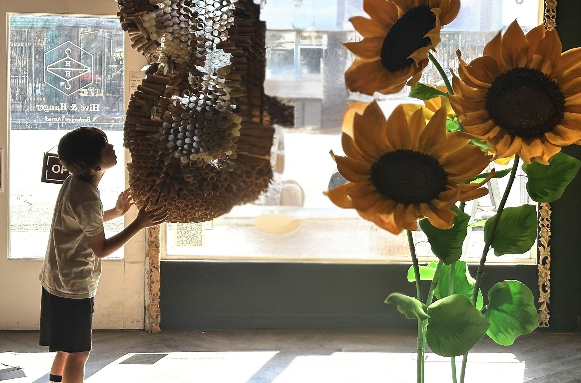A child stands by a large window indoors, touching a large hanging woven beehive sculpture. In the foreground, oversized sunflower decorations with green leaves are visible against a bright window.